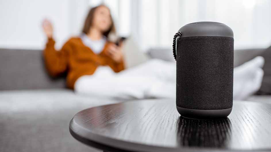 Een zwarte luidspreker op een ronde tafel. Op de achtergrond zit een persoon op een grijze bank met witte kussens.