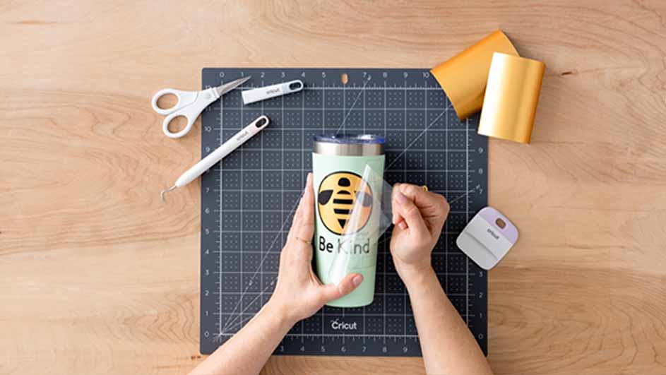 Een lichtgroene beker met een bij-ontwerp wordt beplakt op een grijze snijmat, met gereedschap, schaar en goudkleurig vinyl.