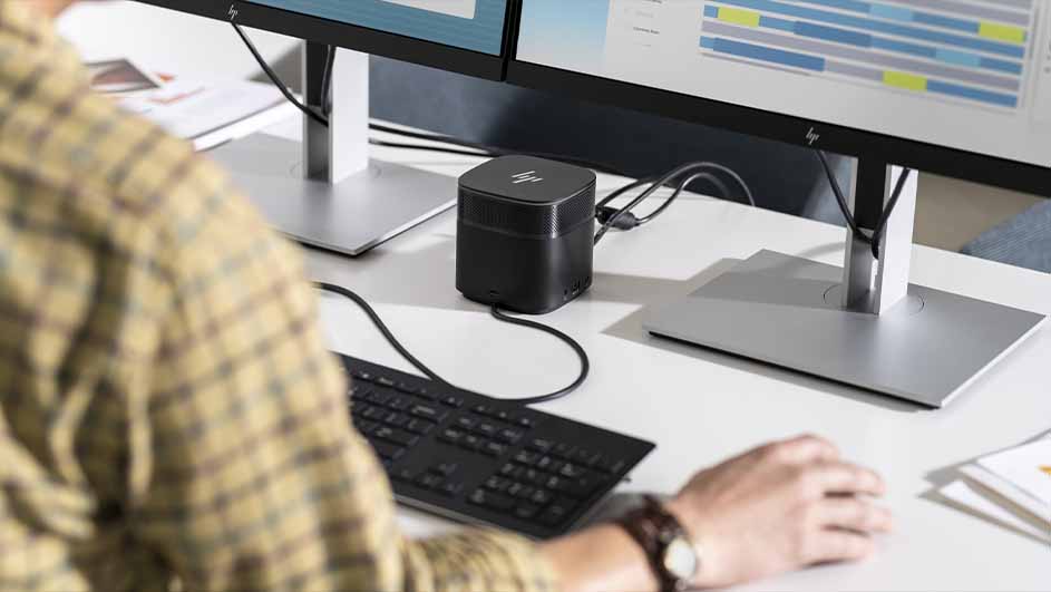 Een bureau met twee monitoren, een zwart toetsenbord en een kleine zwarte computer. Iemand zit aan het bureau met een horloge om.