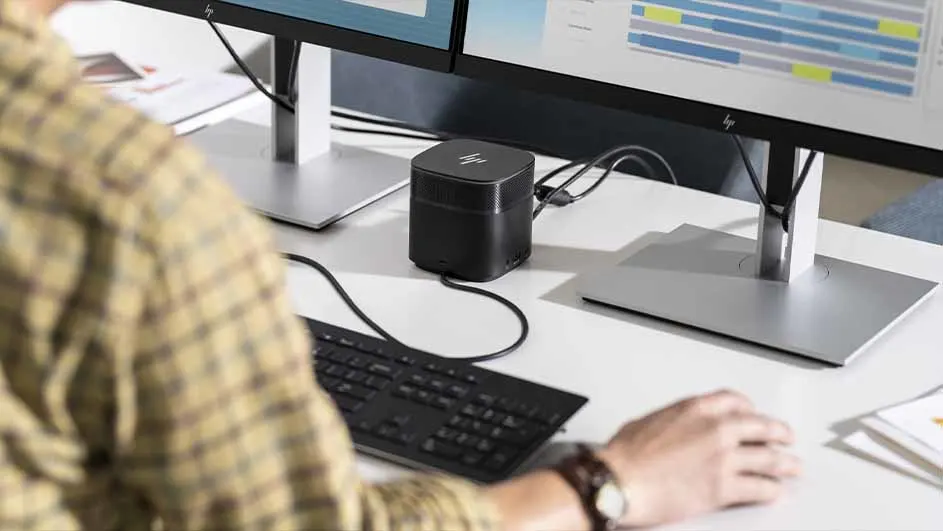 Een bureau met twee monitoren, een zwart toetsenbord en een kleine zwarte computer. Iemand zit aan het bureau met een horloge om.
