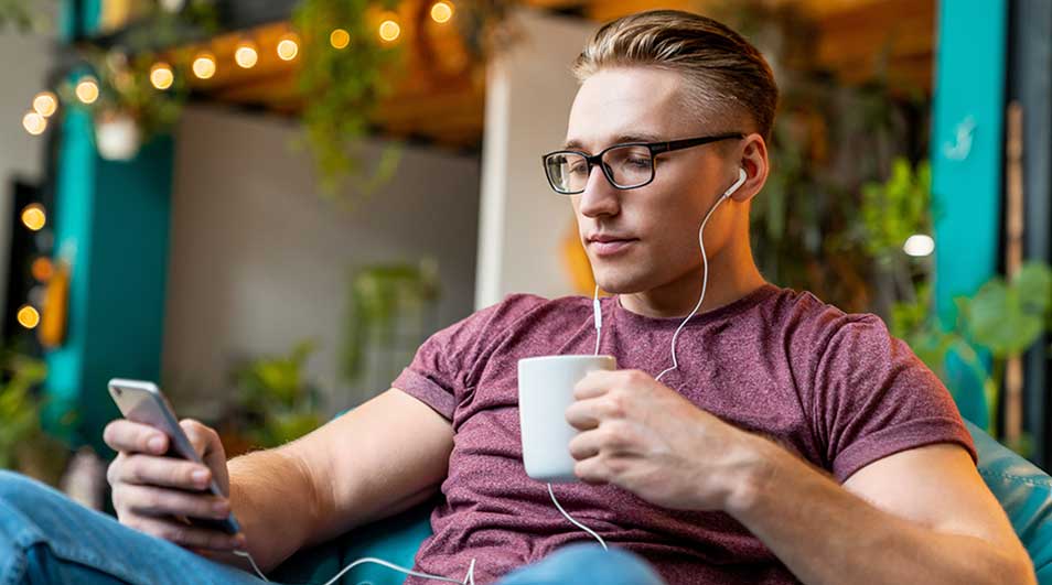 Jonge man met bril zit ontspannen in een café, luistert met bedrade oordopjes naar zijn telefoon terwijl hij een warme mok vasthoudt.
