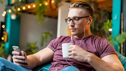 Jonge man met bril zit ontspannen in een café, luistert met bedrade oordopjes naar zijn telefoon terwijl hij een warme mok vasthoudt.