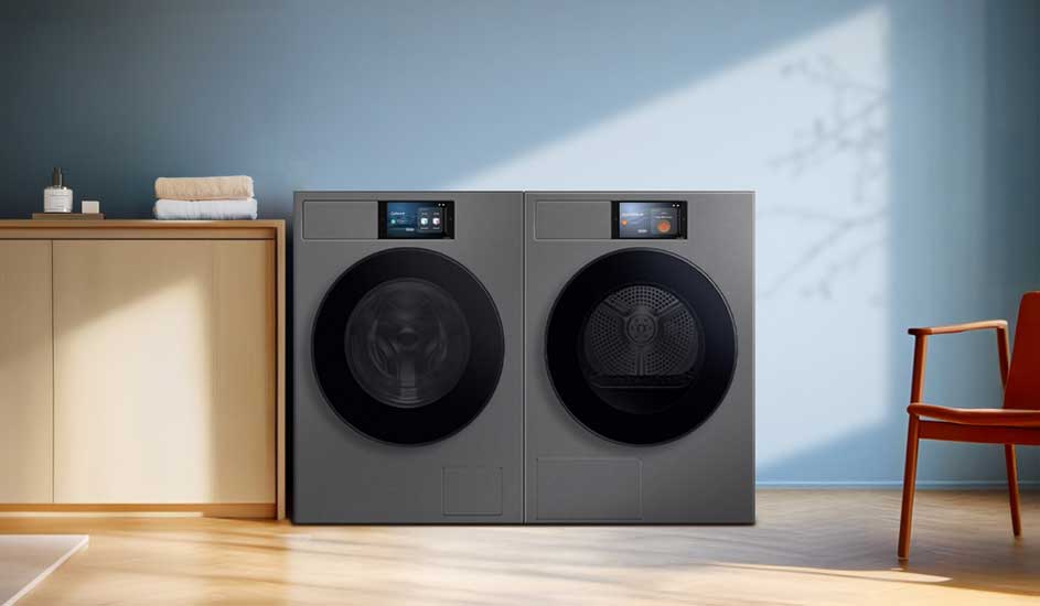 Moderne wasruimte met twee grijze frontloaders naast elkaar, bovenop digitale displays; links kast met handdoeken, rechts een houten stoel.