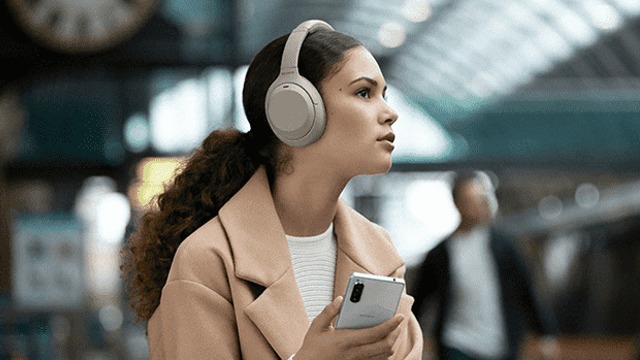 Woman with beige headphones and coat holds a smartphone. Striped white shirt is visible. Background is blurry with clock and people.