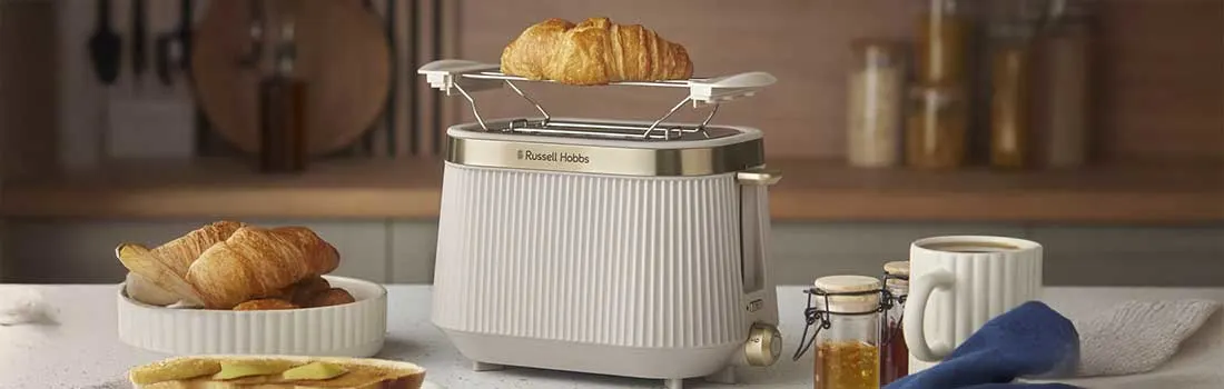 A stylish white Russell Hobbs toaster with warming rack holds a croissant, surrounded by toast with butter, jam jars, a white mug of coffee and breakfast dishes.