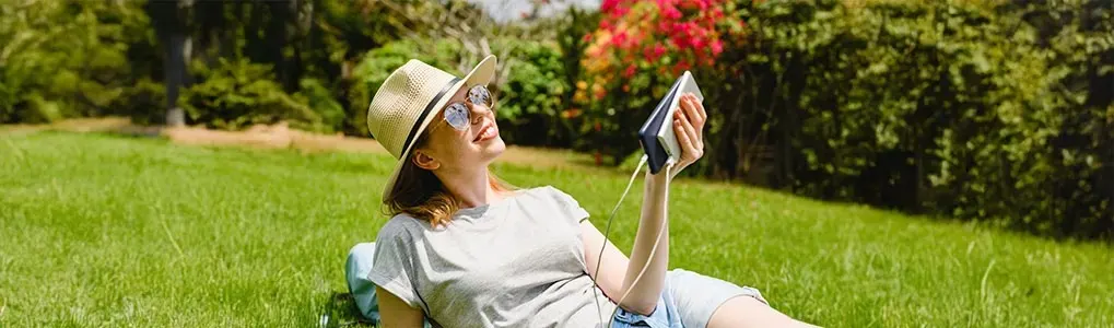 Person with hat and glasses lying on grass, holding a phone connected to a charger, against a backdrop of trees and flowers.