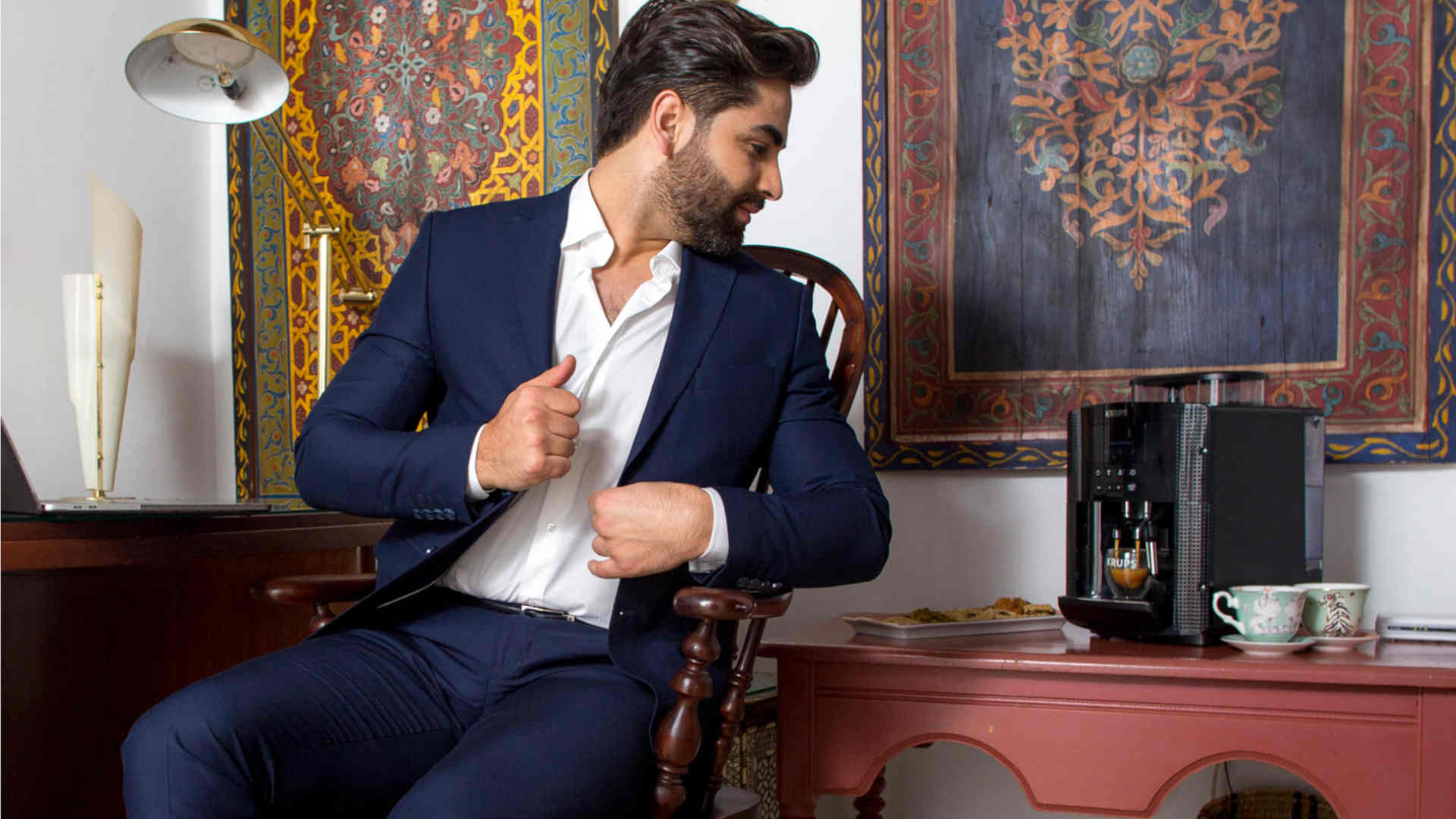 Een man in een blauw pak zit op een houten stoel, naast een koffiezetapparaat op een roodachtig bruine tafel.