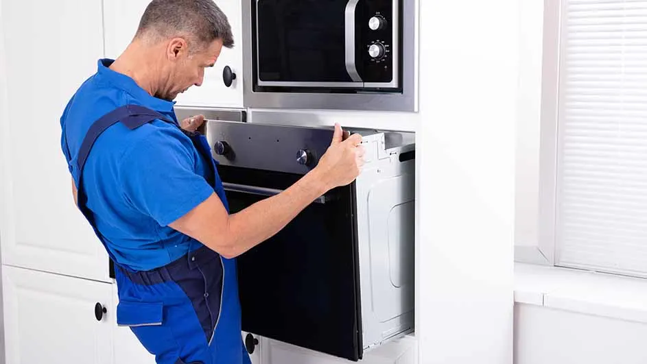 Een man in blauwe werkkleding installeert een oven in een witte keukenkast, met een magnetron erboven.