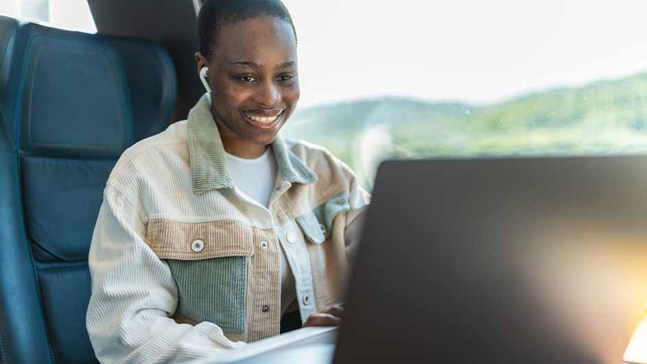 Een vrouw lacht en gebruikt een laptop in een trein. Ze draagt oortjes en een jas met knopen. Buiten is groen te zien.