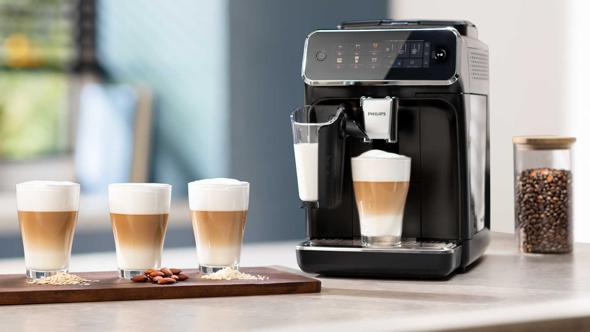Een zwarte Philips-koffiemachine naast vier glazen latte macchiato op een houten plank, met een pot koffiebonen.