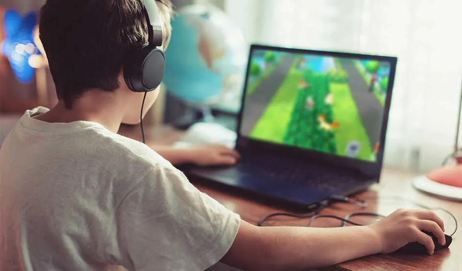 A boy wearing headphones is playing a colourful game on a laptop at a desk. The laptop brand is not visible in the image.