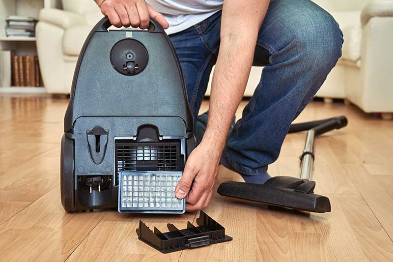 Person kneeling on a wooden floor, replacing the filter at the back of a canister vacuum cleaner.