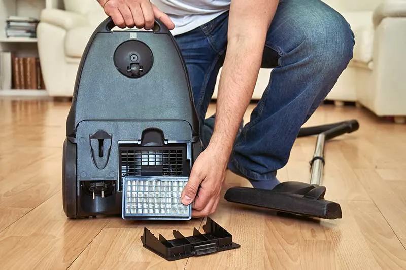 Person kneeling on a wooden floor, replacing the filter at the back of a canister vacuum cleaner.