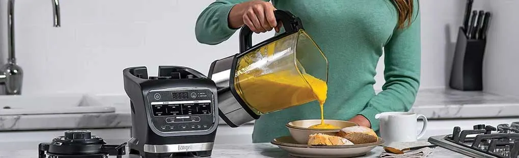 Een vrouw in een keuken giet gele soep uit een blenderkan in een kom; op het aanrecht staan blenderbasis, brood en een kopje.