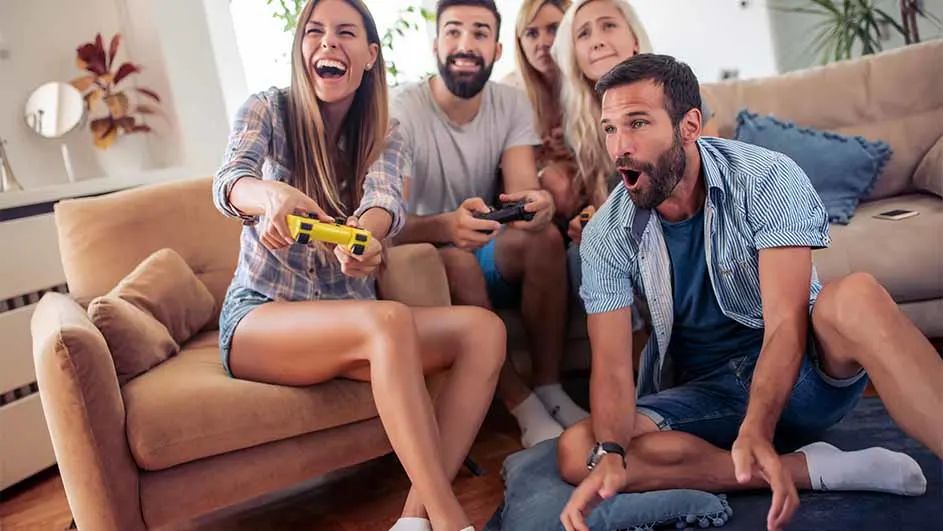 Vijf mensen spelen videospelletjes in een woonkamer, zittend op een beige bank en vloerkussen. Een vrouw lacht met een gele controller.