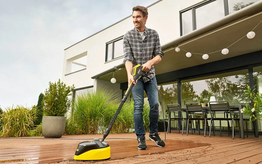 Een glimlachende man in geruit overhemd reinigt zijn houten terras met een gele schijfborstel voor zijn moderne huis met glazen wanden en verlichting.