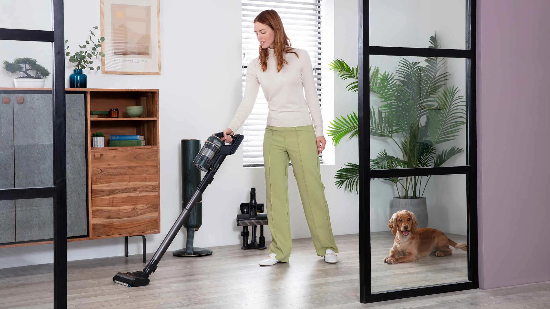 Een vrouw stofzuigt in een kamer met houten vloeren, een kast, een plant en een hond ligt in de deuropening.