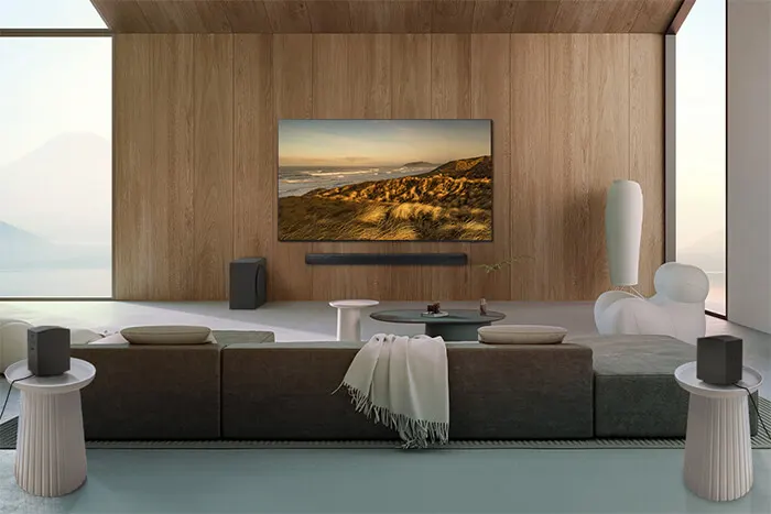 Woonkamer met houten wand, tv, modulaire bank met kussens en kleed, bijzettafels en speakers. Een groot raam laat natuurlijk licht binnen.