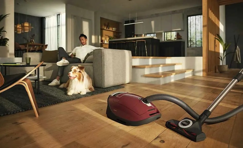 A red Miele cylinder vacuum cleaner is on a wooden floor in a modern living room, with a dog lying on the rug next to the sofa.