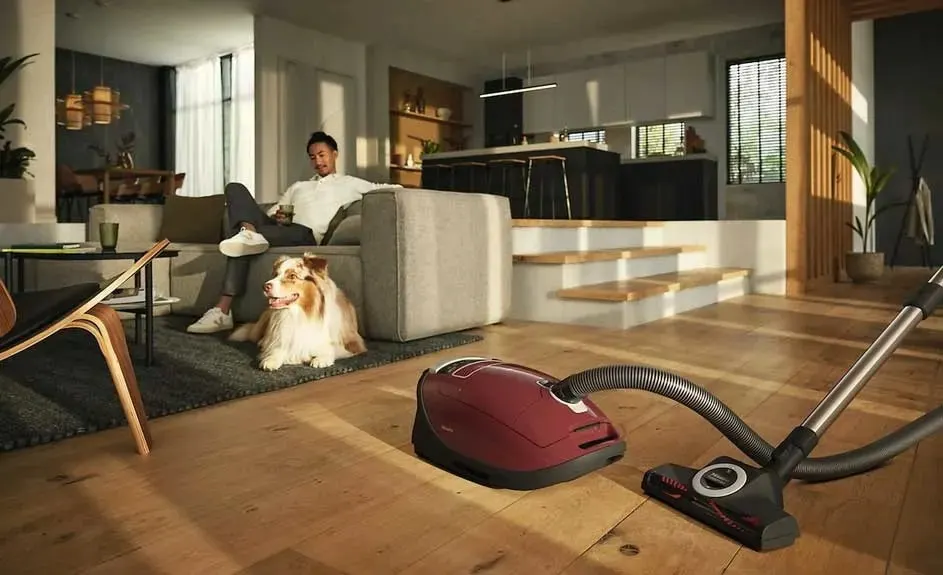 A red Miele cylinder vacuum cleaner is on a wooden floor in a modern living room, with a dog lying on the rug next to the sofa.