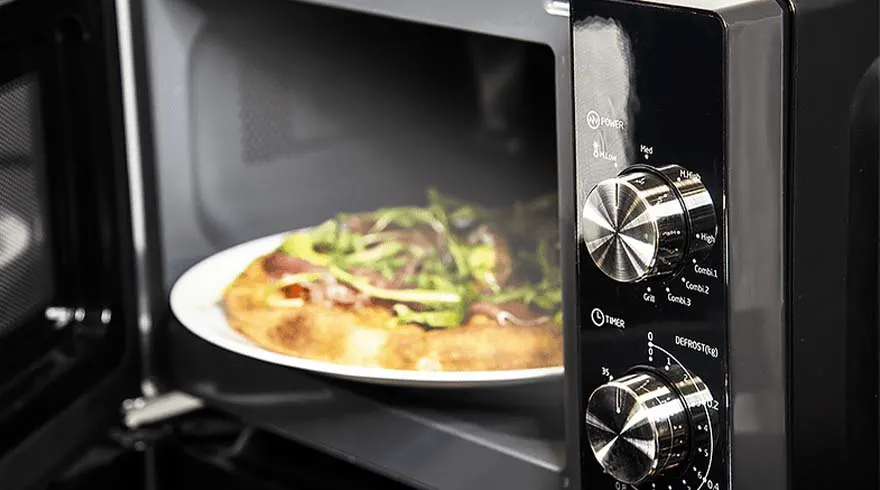 Pizza op een wit bord in een open, zwarte magnetron, met bedieningsknoppen aan de rechterkant.