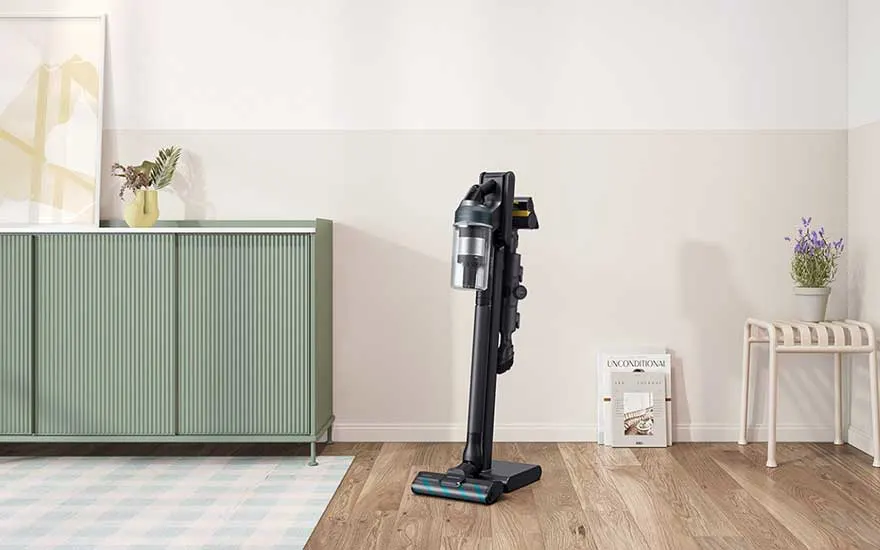 A black cordless vacuum stands upright in a tidy living room, positioned beside a green sideboard, magazines and potted plants.