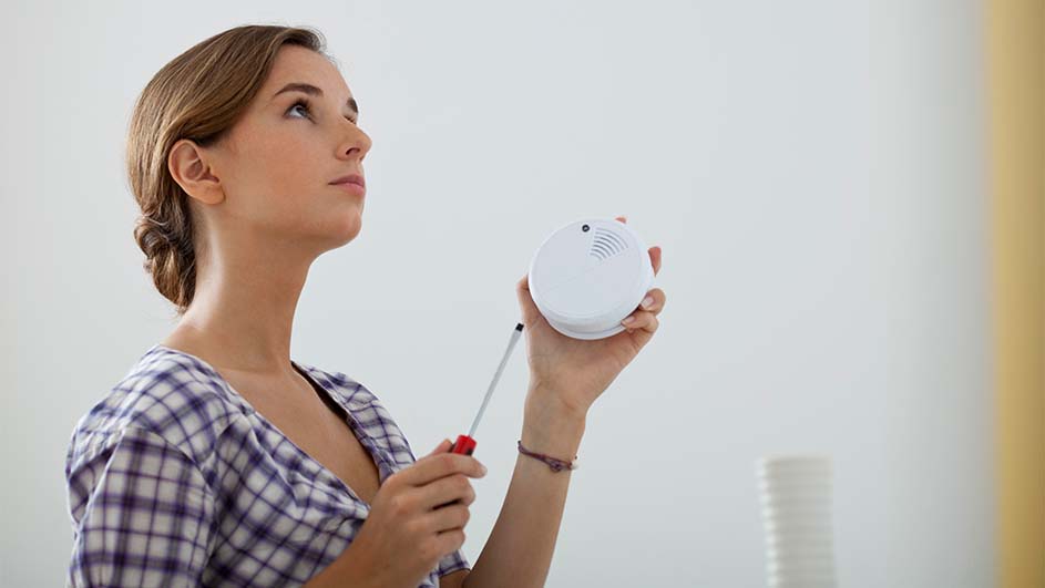 Vrouw met een schroevendraaier en een rookmelder in haar handen, kijkend naar het plafond in een kamer met witte muren.