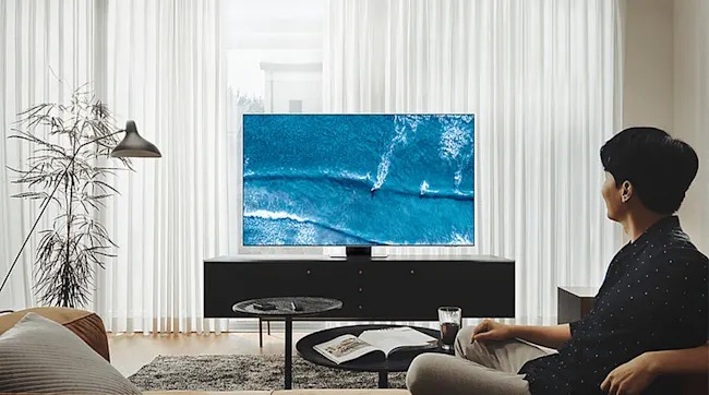 Een man zit op de bank en kijkt naar een tv met een beeld van zee en surfers. De woonkamer is licht met moderne meubels en veel daglicht.
