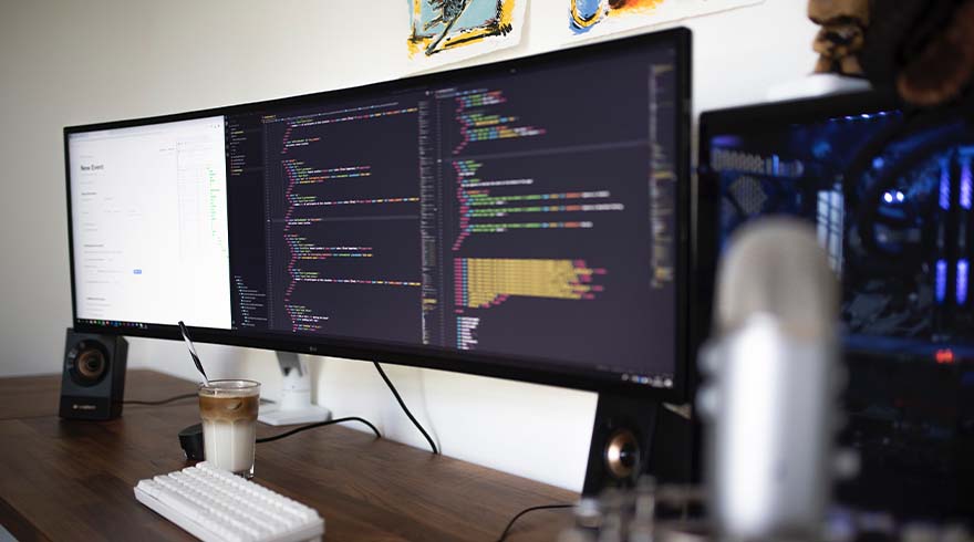 Een bureau met een toetsenbord, drankje en drie schermen met code en een programma. Luidsprekers aan de zijkanten.