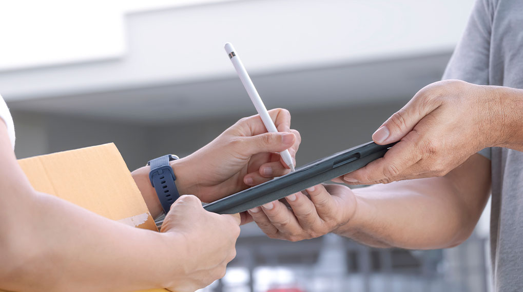 Een persoon tekent met een witte stylus op een tablet. Een andere persoon houdt de tablet vast en een pakje.