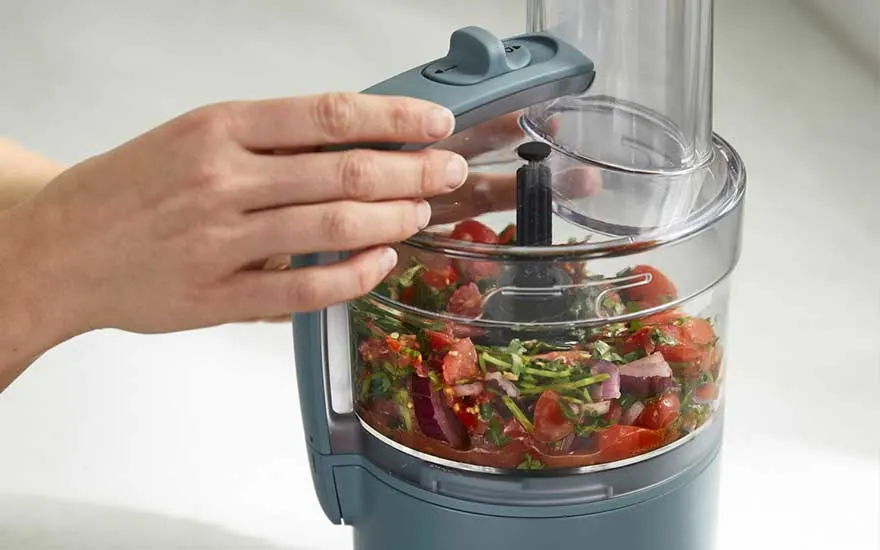 Een hand sluit de deksel van een compacte keukenmachine, waarin fijngesneden tomaten, ui en groene kruiden klaarstaan om snel verder te worden gehakt.