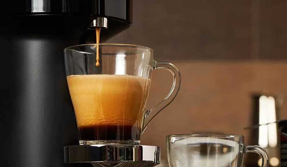 A Senseo coffee machine pours fresh espresso into a glass mug, creating a rich crema; another empty mug sits beside it on a kitchen counter. Brand: Senseo.