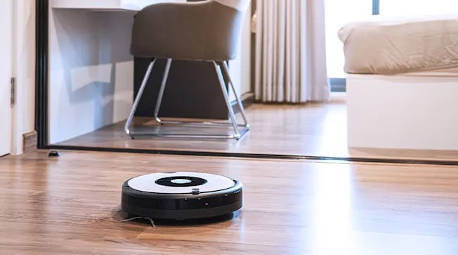 Een ronde, zwarte robotstofzuiger staat op een houten vloer in een kamer met een bureau en een bed.