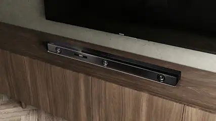 Een zwarte soundbar staat op een houten tv-meubel onder een zwart tv-scherm aan de muur.