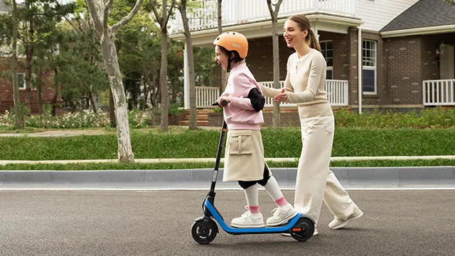 Een kind met helm rijdt op een blauwe scooter, begeleid door een volwassene op een weg met huizen en groen.