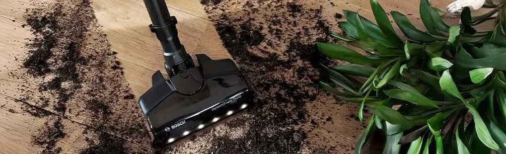 Een zwarte stofzuiger zuigt aarde op van een houten vloer naast een groene plant.