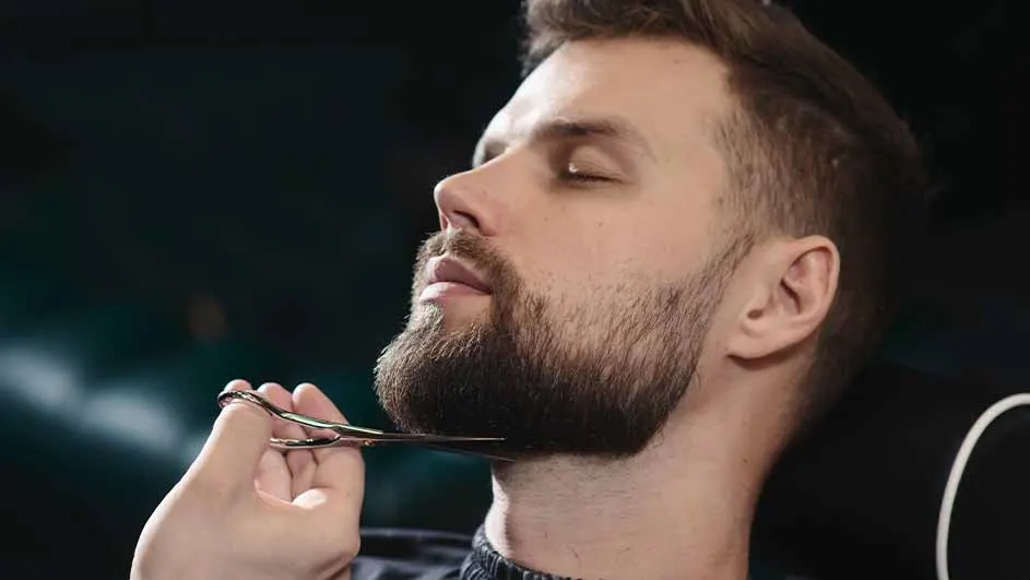 Een man met een baard krijgt zijn baard geknipt met een zilveren schaar tegen een donkere achtergrond in een kapperszaak.