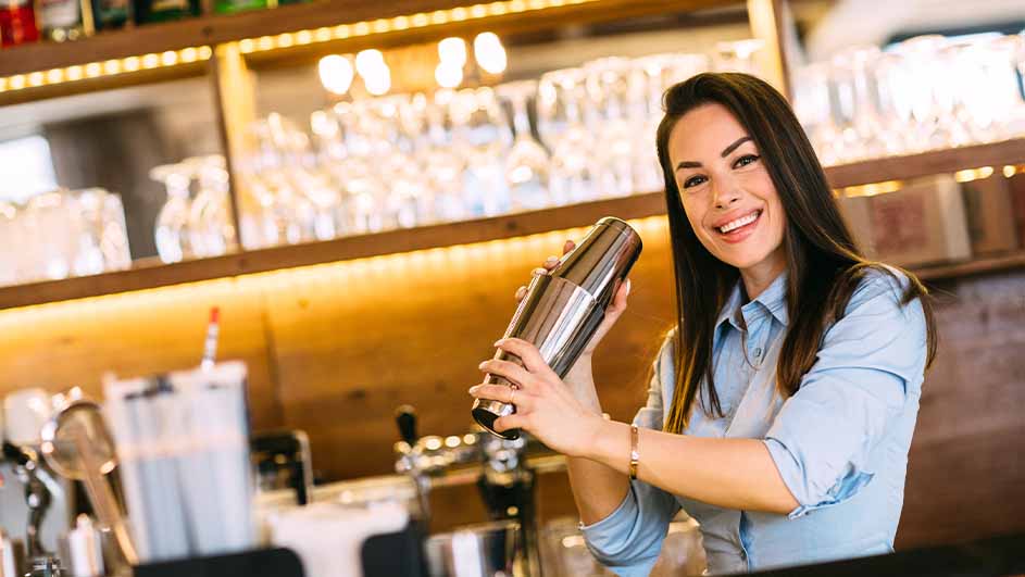 Een vrouw in een blauw hemd schudt een cocktailshaker in een bar. Achter haar staan glazen en flessen op houten planken.