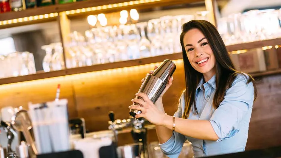 Een vrouw in een blauw hemd schudt een cocktailshaker in een bar. Achter haar staan glazen en flessen op houten planken.