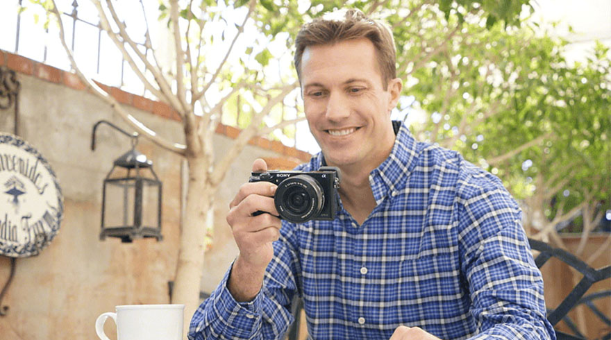 Een man met een geruit blauw overhemd houdt een zwarte camera vast. Achter hem staan een lantaarn, een bord en groen gebladerte.