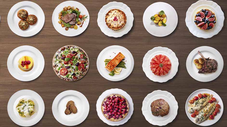 Diverse gerechten op witte borden, gerangschikt op een houten tafel. Variërend van gebak tot vis, vlees en fruit.