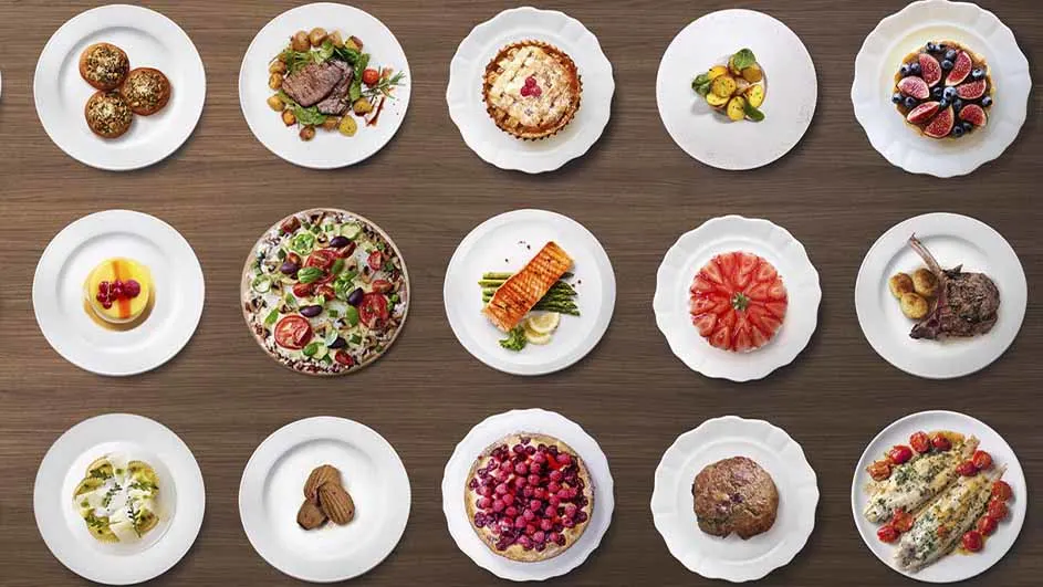 Diverse gerechten op witte borden, gerangschikt op een houten tafel. Variërend van gebak tot vis, vlees en fruit.