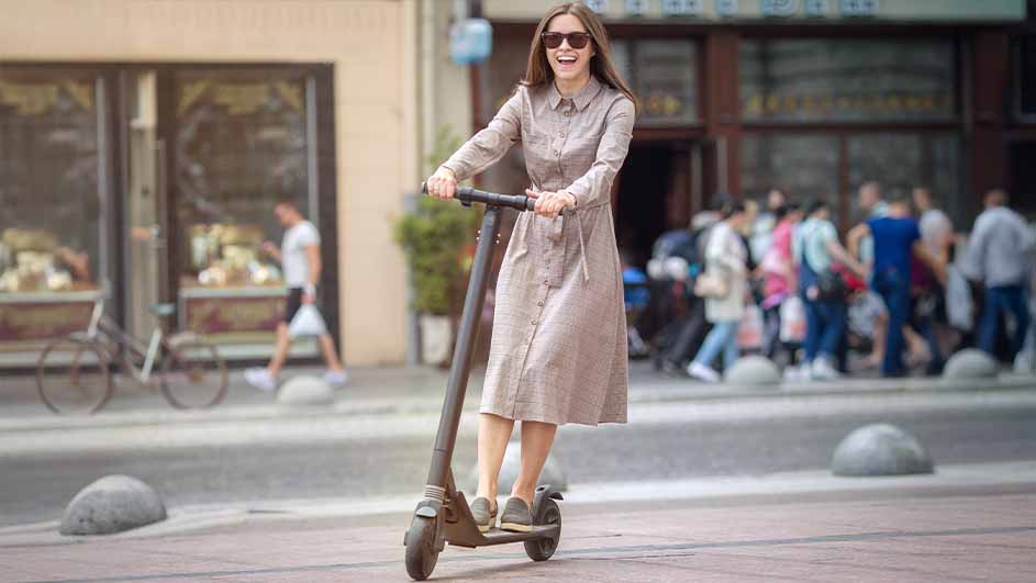A person in a dress and sunglasses rides an electric scooter on a city street with pedestrians and buildings in the background.