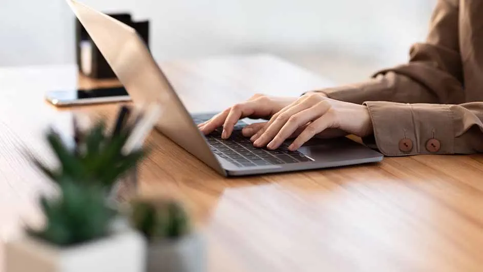 Persoon typt op een laptop op een houten bureau. Een telefoon en een plant staan op het bureau.