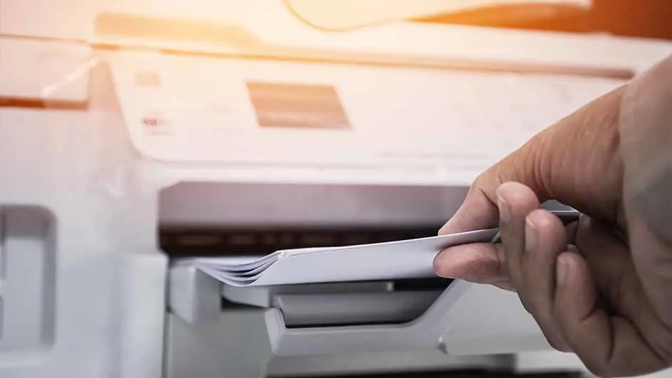 Een hand plaatst papier in een witte printer. De printer staat op een vlakke ondergrond.