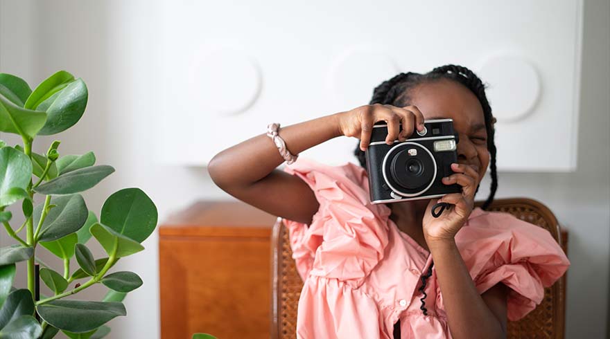 Een kind zit op een stoel en houdt een zwarte camera voor haar gezicht. Op de achtergrond een witte muur en een houten kast. Links een groene plant.