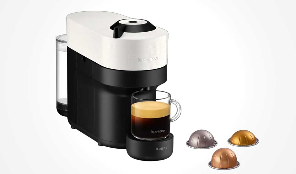 Een compacte capsulekoffiemachine zet koffie in een glazen kopje, met waterreservoir achteraan en drie capsules op de lichte ondergrond.