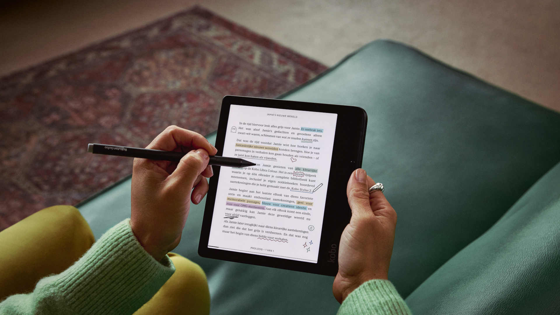 Person holds a Kobo eReader with stylus, highlighting text. Reader rests on a green surface. A hand wears a ring.