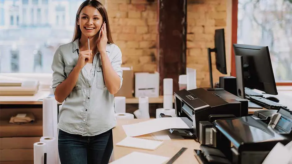 Vrouw aan de telefoon in kantooromgeving met printers, papier en computers op een bureau voor een bakstenen muur.