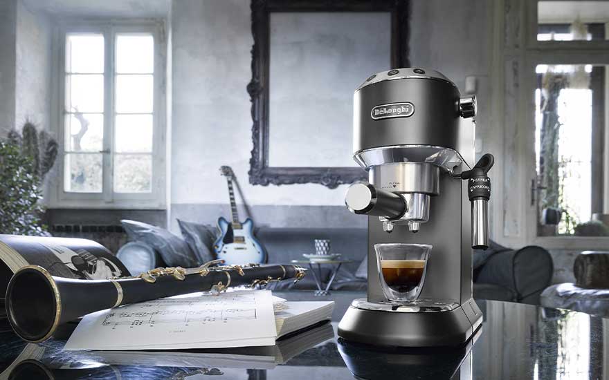 A compact De’Longhi espresso machine sits on a glossy table beside sheet music and a clarinet, with a guitar in the background.